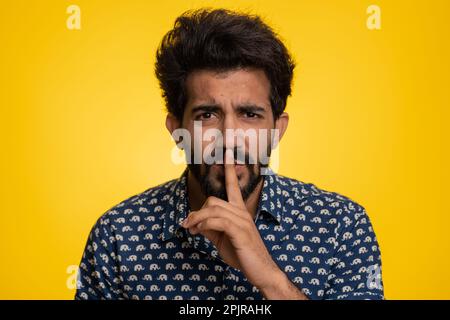 Shh be quiet please. Young indian man presses index finger to lips makes silence gesture sign do not tells secret, stop gossip. Handsome bearded hindu guy isolated alone on yellow studio background Stock Photo