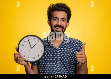 It is your time. Excited happy young indian man showing time on wall office clock, ok, thumb up, approve, pointing finger at camera. Handsome hindu guy isolated on studio yellow background indoors Stock Photo