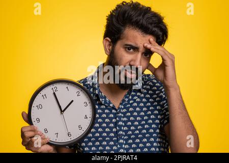 Young indian man with anxiety checking time on clock, running late to work, being in delay, deadline. Hindu guy looking at hour, minutes, worrying to be punctual isolated on studio yellow background Stock Photo