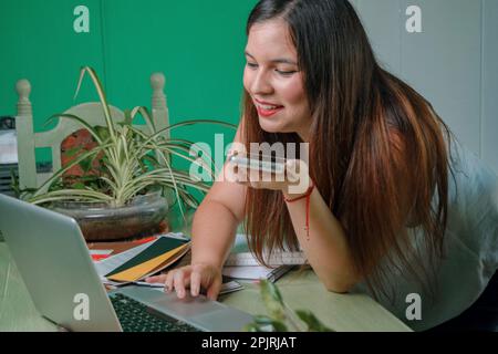 young hispanic latin business woman working at home using her laptop and smiling sending a voice note on the phone. Stock Photo