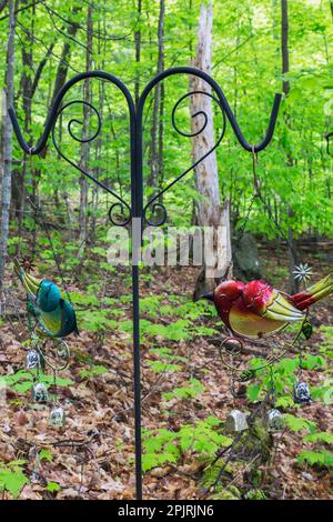 Colourful painted metal bird wind chimes hung on black wrought iron stand in forest of deciduous trees in backyard garden in spring. Stock Photo