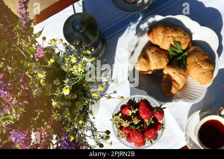 Beautiful bouquet of wildflowers, ripe strawberries, tea and croissants on table outdoors, flat lay Stock Photo