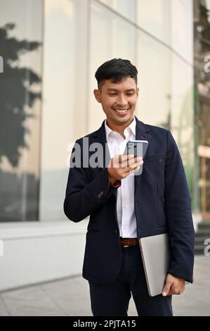 Portrait, Handsome millennial Asian businessman in a formal business suit texts with someone on his smartphone while walking in a city. Stock Photo