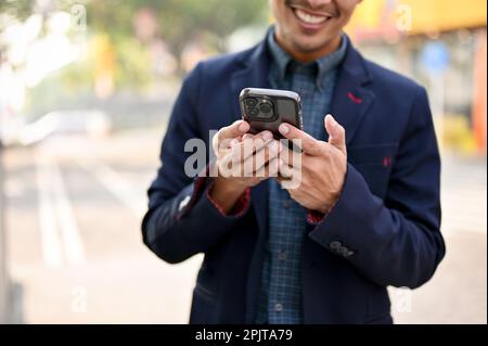 Close-up image of smiling and handsome millennial Asian businessman using his smartphone while walking in a city. Stock Photo