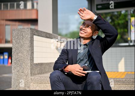 Smiling millennial Asian businessman resting on a stairway in front of the metro train station in a city on a sunny day. Stock Photo