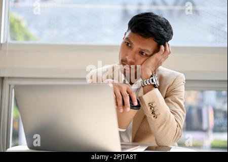 Bored and tired millennial Asian businessman working on his project on laptop, remote working at cafe. Stock Photo