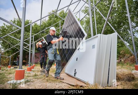 Workers carrying solar panels for installing in field. European men wearing workwear and helmets. Photo-voltaic collection of modules as a PV panel. Array as a system of photo-voltaic panels. Stock Photo