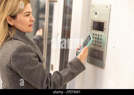 Young stylish woman getting access to the building by attaching smartphone to intercom. Concept of modern security technologies for access and smart Stock Photo