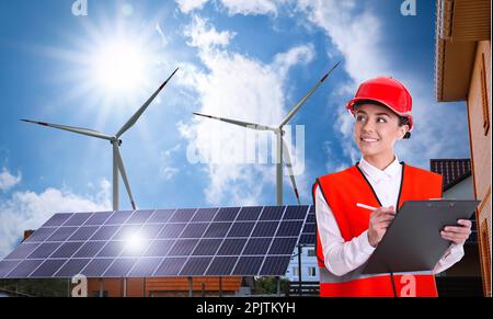 Industrial engineer in uniform and view of wind energy turbines near house with installed solar panels on roof Stock Photo