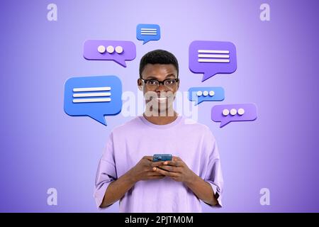 Cheerful young African American man in glasses using smartphone over purple background with speech bubbles. Concept of social media and chatting Stock Photo