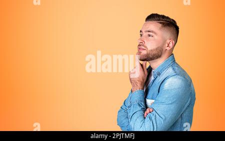 Pensive young handsome man with hand on chin, profile portrait making up plans, orange empty copy space background. Concept of thoughts and idea Stock Photo