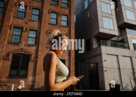 Fitness girl in sport outfit wearing headphones and turning music on smartphone. running in city Stock Photo