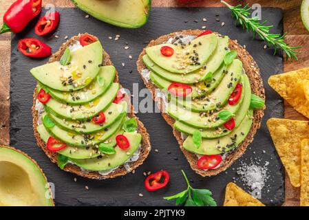 Avocado toasts - bread with avocado slices, pieces of red pepper and sesame on black stone board. Stock Photo