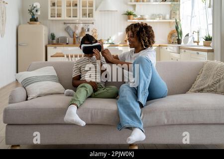 Mother helping son to wear new device virtual reality glasses while sitting together on couch Stock Photo