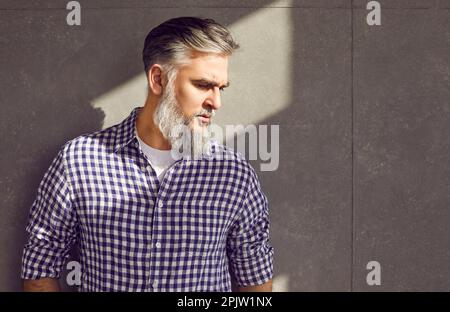 Handsome senior bearded male model in casual shirt leaning on grey wall background Stock Photo