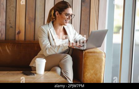business woman using a laptop while working in a cafe. Professional woman doing remote work in a coffee shop. Happy female freelancer sitting on a cou Stock Photo