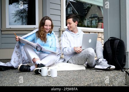 schoolchildren teenagers on doorstep at home doing homework boy in hands of laptop girl has big white folder two cups of tea other embarrassment first relationship friendship between different sexes Stock Photo
