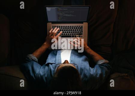 Young coder in eyeglasses is working remotely from home late at night, looking on display, typing code. Hard worker, programmer working overtime, taking work at home, has deadline.  Stock Photo