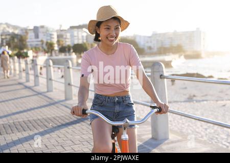 Happy asian woman riding bike along promenade by the sea Stock Photo