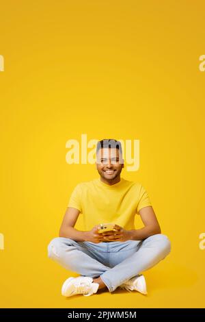 Smiling indian man using smartphone sitting on the floor in relaxed pose, isolated on yellow backdrop, carefree serene male student chatting, spends time in social networks, scrolling news feed Stock Photo