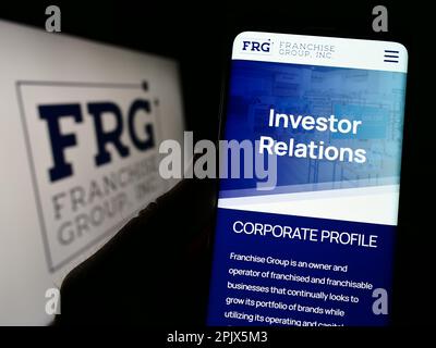 Person holding cellphone with webpage of US company Franchise Group Inc. (FRG) on screen in front of logo. Focus on center of phone display. Stock Photo