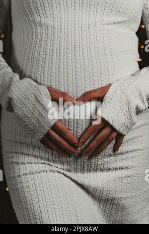 A pregnant woman in a white dress gesturing with her hands to form a heart shape over her belly Stock Photo