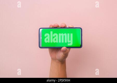 Human hand holding mobile smartphone with green screen in horizontal position isolated on pink background. Stock Photo