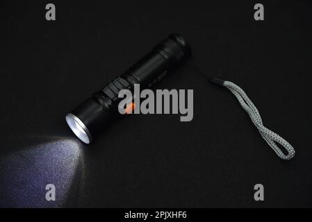 Metal black flashlight on a black background. Burning pocket flashlight close-up. Lighting device. Stock Photo