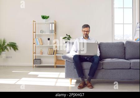 Mature serious man is working on laptop at home office sitting on couch, remote dostance work. Stock Photo