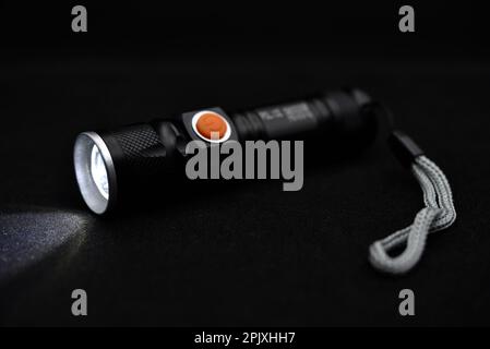 Metal black flashlight on a black background. Burning pocket flashlight close-up. Lighting device. Stock Photo