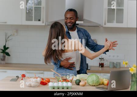 Happy romantic interracial couple dancing in the kitchen while cooking together. African-American man and woman having fun in the kitchen Stock Photo