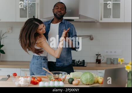 Happy romantic interracial couple dancing in the kitchen while cooking together. African-American man and woman having fun in the kitchen Stock Photo