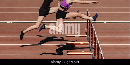 two female athletes running hurdles in athletics competition, hurdling on stadium track, summer sports games Stock Photo