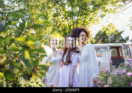 Pretty smiling three teenage girls in light dresses. Female teens of different nationalities by retro minibus. Old car is decorated with macrame panel Stock Photo