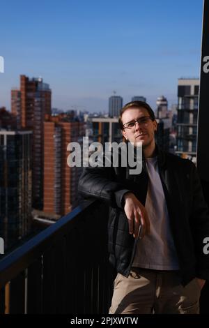 Trendy handsome thoughtful caucasian man in jacket, shirt and glasses leaning on fence of a balcony in a big modern city at home looking in camera and Stock Photo