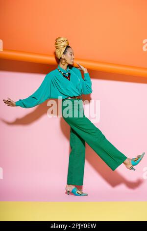 full length of cheerful african american woman in stylish outfit smiling while holding orange rolled wallpaper on pink background Stock Photo