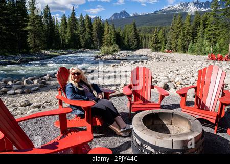 Woman relaxes around a campfire ring sitting on a red adirondack chair, in the Canadian Rockies Stock Photo