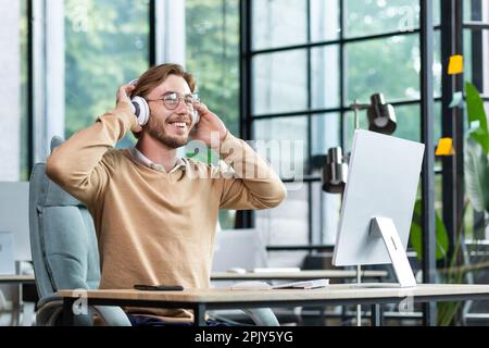 A young male student in headphones studies in the office, campus, at home in front of a laptop, hands up, dances, listens to a lecture. Smiling, resting. Stock Photo