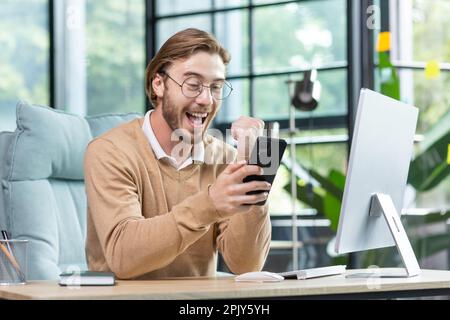Happy young student in glasses sitting in office at home in front of computer, holding phone in hand, showing victory success gesture with hand, celebrating, smiling. Stock Photo