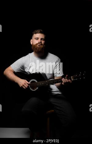 Handsome brunette bearded man musician plays an acoustic guitar in a black room. Ready to play a music. Stock Photo