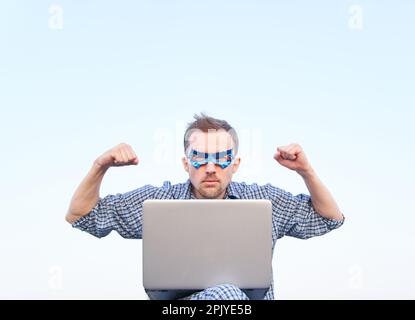 Pumped fists or superpower, serious emotion concept. Caucasian male in nightwear shirt and superhero blue mask using laptop and showing strength gesture. High quality image Stock Photo