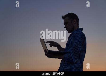 Online meeting concept. Adorable freelancer or IT support male engineer working outdoor using laptop pointing on screen with finger in silhouette at sunset. High quality image Stock Photo