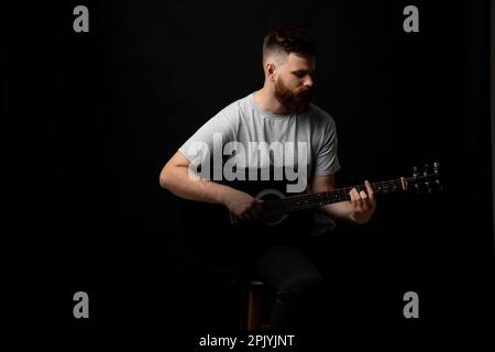 Handsome bearded brutal man musician in a grey t-shirt holding a acoustic guitar on a shoulder on black background. Stock Photo
