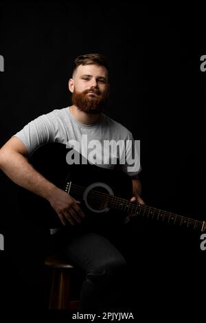 Young bearded guitarist in grey t-shirt holds a guitar and playing a song in a dark room. Stock Photo