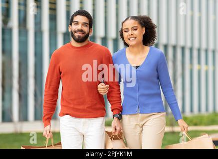 Happy Multicultural Buyers Couple Holding Paper Shopping Bags Walking Outdoors Stock Photo