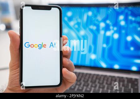 A mans hand holding a smartphone with Google AI neural network logo on the screen and a laptop displaying a neuronet on the splash screen in the background. March 2023, Prague, Czech Republic  Stock Photo