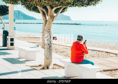 Unrecognizable eldery woman recording voice messages using smartphone online, calling or listen voice message. Woman relaxing, sitting on bench Stock Photo
