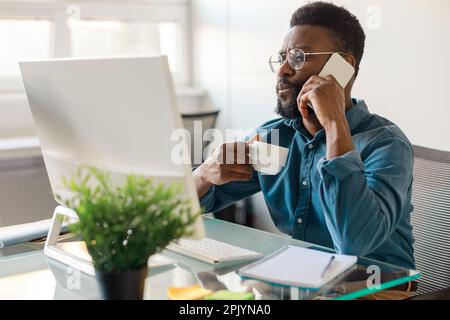 Remote business. African american male ceo using computer, having corporate phone conversation call and drinking coffee Stock Photo