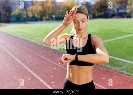 Tired and upset mature female athlete checking heart rate on smart watch fitness bracelet, runner in stadium after active exercise and jogging on sunny day, blonde woman in sportswear. Stock Photo