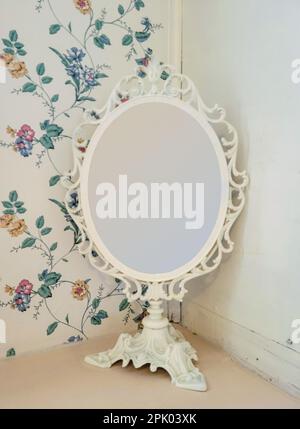 Old-fashioned ivory ornate mirror on a pedestal with retro floral wallpaper background Stock Photo